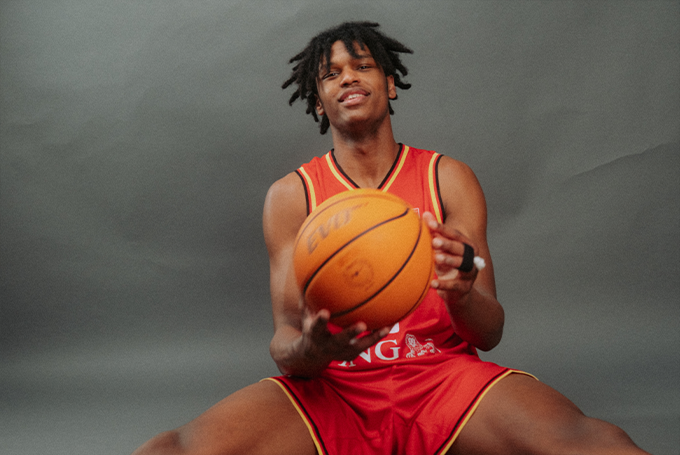 Dieses Bild zeigt einen jungen Basketballspieler, der in einem roten Trikot entspannt sitzt und einen Basketball in den Händen hält.